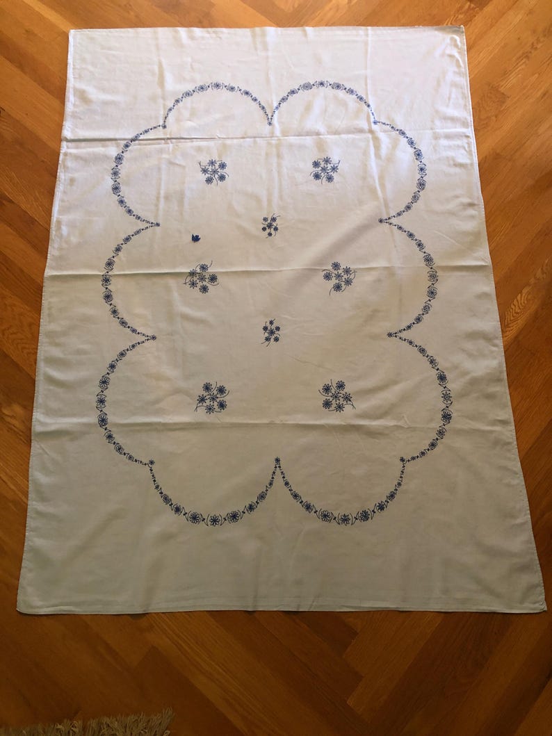 May include: A white tablecloth with a large, scalloped border and floral embroidery in blue. The embroidery features clusters of small flowers and a delicate, repeating pattern along the edge. The tablecloth is rectangular and appears to be made of a soft fabric.