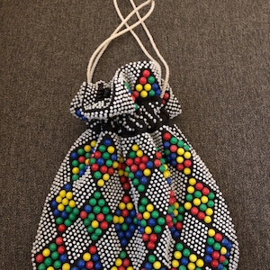 May include: A drawstring pouch covered in small white beads featuring a geometric diamond pattern. The diamonds are filled with clusters of red, yellow, green, and blue beads. The bag includes two white cord handles that cinch the top opening closed.