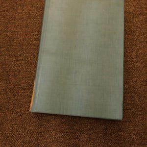 May include: A hardcover book with a light blue cover. The book's cover is slightly worn, with a visible crease near the spine. The book is resting on a brown, textured surface.