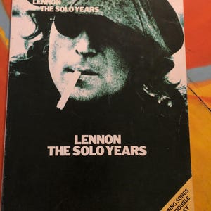 May include: A black and white photo of John Lennon on the cover of a music book titled "Lennon The Solo Years: Easy Guitar". The book features songs from the album "Double Fantasy".