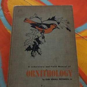 May include: A vintage book titled "A Laboratory and Field Manual of Ornithology" by Olin Sewall Pettingill, Jr. The cover features a black and orange bird perched on a branch with leaves. The book has a gray cover.