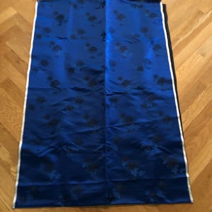 May include: A rectangular piece of royal blue fabric featuring a repeating black floral pattern. The fabric has a smooth, satin-like finish and is bordered by a thin white stripe along the vertical edges, with raw, unfinished edges at the top.