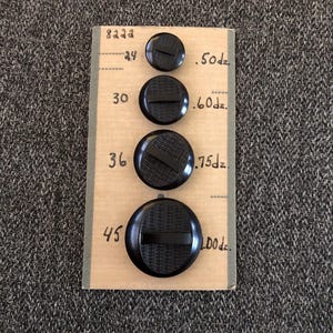 May include: Four black buttons of varying sizes are displayed on a tan card with handwritten measurements. The buttons have a textured center and a horizontal bar across the middle. The card has the numbers 24, 30, 36, and 45.