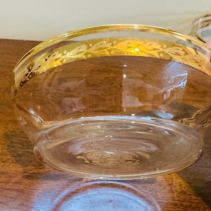 May include: Clear glass bowl with a gold rim featuring a decorative leaf pattern. The bowl has a rounded shape and sits on a wooden surface. The gold rim adds a touch of elegance to the simple design.