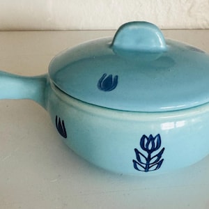 Può includere: Una zuppiera in ceramica azzurra con coperchio e manico. La zuppiera è decorata con motivi floreali blu scuro. Il manico è sul lato sinistro. Il coperchio ha un piccolo pomello.