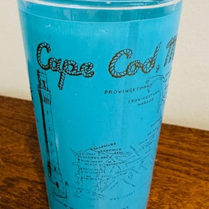 May include: A clear glass with a light blue background featuring the words "Cape Cod" in a rope-like font. The glass has a map of the Cape Cod area with landmarks and town names in black.