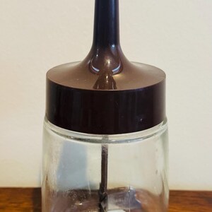 May include: A vintage manual food chopper with a clear glass jar, a brown plastic lid and handle, and a stainless steel blade. The white handle is at the top. The chopper is on a wooden surface.