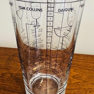 May include: Clear cocktail mixing glass with black printed recipes for Tom Collins and Daiquiri cocktails. The glass features measurement markings and ingredient lists, perfect for home bartenders. The glass is approximately 6 inches tall.