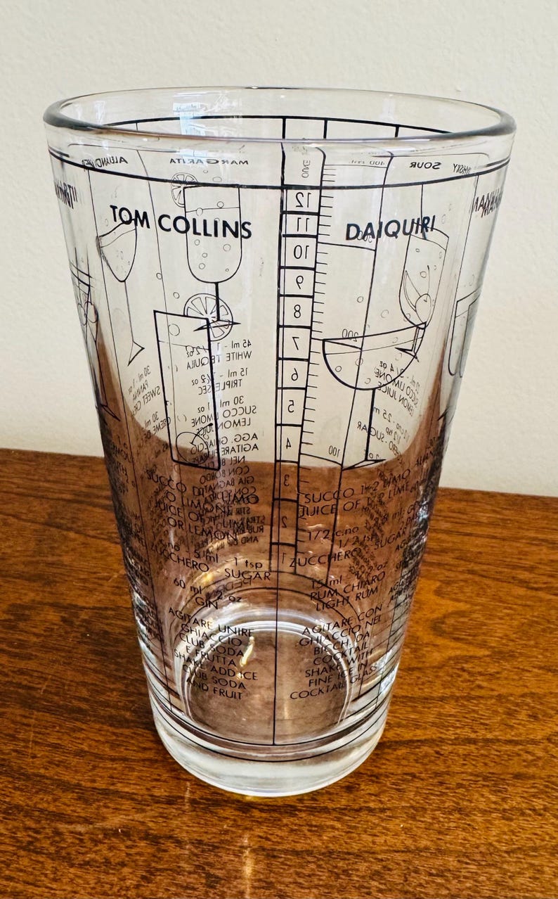 May include: Clear cocktail mixing glass with black printed recipes for Tom Collins and Daiquiri cocktails. The glass features measurement markings and ingredient lists, perfect for home bartenders. The glass is approximately 6 inches tall.