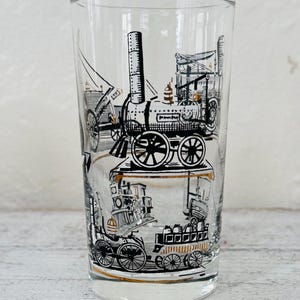 May include: Clear glass tumbler featuring black and gold illustrations of vintage trains. The design includes detailed depictions of steam engines and carriages, creating a nostalgic aesthetic. The glass is cylindrical and sits on a light-coloured wooden surface.