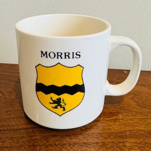 Puede incluir: Taza de cerámica blanca con el nombre "MORRIS" impreso sobre un escudo amarillo y negro. El escudo presenta una línea negra ondulada y una pequeña figura negra. La taza tiene un asa curva.