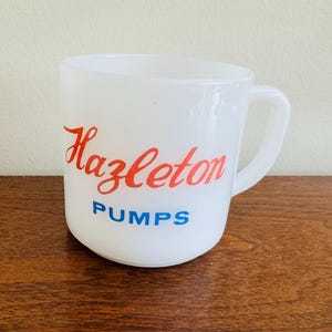 Puede incluir: Taza blanca con asa. La taza presenta el nombre "Hazleton" en escritura roja y la palabra "PUMPS" en letras azules.