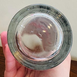 May include: A clear glass with a gray rim and a slightly textured base. The glass is held by a hand, and the bottom of the glass is visible, showing a circular shape.