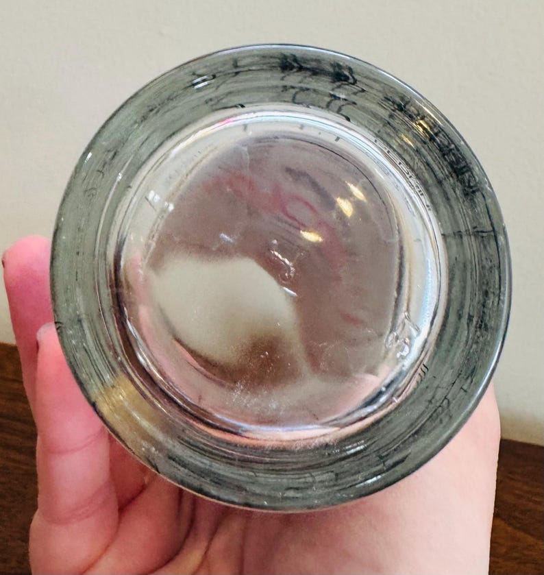 May include: A clear glass with a gray rim and a slightly textured base. The glass is held by a hand, and the bottom of the glass is visible, showing a circular shape.