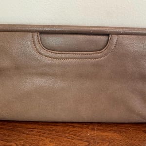 Puede incluir: Un bolso de mano rectangular de color topo con asa. El bolso está hecho de un material liso, posiblemente cuero o un tejido similar al cuero. El asa tiene un diseño curvo y empotrado. Los bordes están cuidadosamente cosidos.