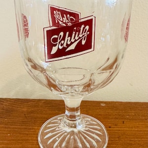 Puede incluir: Copa de cerveza de cristal transparente con pie y cuenco ancho. La copa presenta la palabra "Schlitz" en escritura roja dentro de un rectángulo rojo. Hay gráficos rojos adicionales en el cristal. La copa está sobre una superficie de madera.