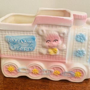 Könnte beinhalten: Ein Keramik-Zug-Pflanzgefäß in Pastellrosa und Weiß mit blauen und gelben Akzenten. Die Worte "BABY LAND EXPRESS" sind auf der Seite zu sehen. Der Zug zeigt ein lächelndes Babygesicht und Blumendetails.