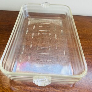 Peut inclure: Un plat de cuisson rectangulaire en verre transparent avec des coins arrondis et des poignées intégrées aux extrémités courtes. La base présente un motif texturé de lignes horizontales et verticales en relief, avec des côtés nervurés.