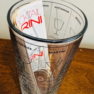 May include: A clear glass cocktail shaker with a black rim and printed cocktail recipes. The shaker contains a stack of recipe cards with the word "COCKTAIL RINI" in red. The shaker has measurements and cocktail names printed on the glass.