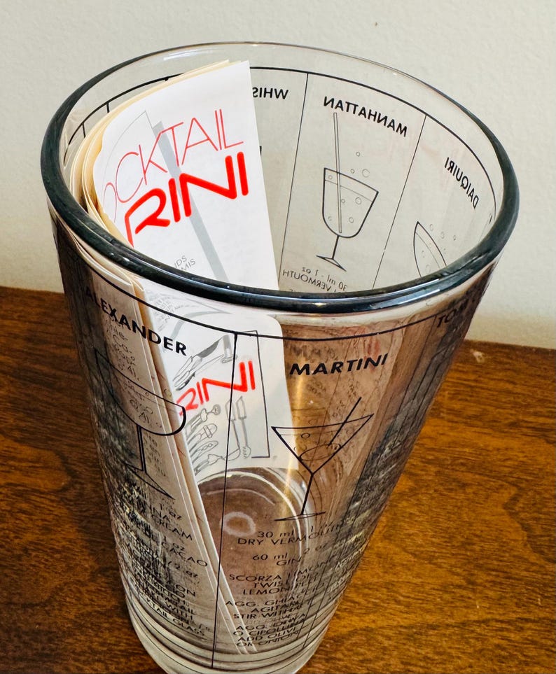 May include: A clear glass cocktail shaker with a black rim and printed cocktail recipes. The shaker contains a stack of recipe cards with the word "COCKTAIL RINI" in red. The shaker has measurements and cocktail names printed on the glass.