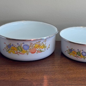 May include: Set of four white enamel bowls with a floral and fruit design. Each bowl has a silver rim and a decorative pattern of yellow, blue, and orange flowers and fruit. The bowls are nested on a wooden surface.
