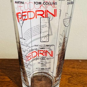 May include: A clear glass cocktail shaker with a black and red design. The shaker features cocktail recipes, including the "Pedrini" and "Tom Collins." Measurements are in ounces and milliliters. The glass is on a wooden surface.