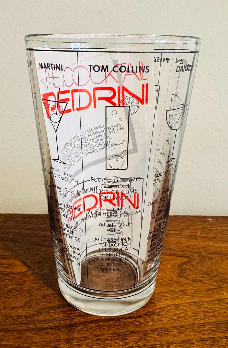 May include: A clear glass cocktail shaker with a black and red design. The shaker features cocktail recipes, including the "Pedrini" and "Tom Collins." Measurements are in ounces and milliliters. The glass is on a wooden surface.