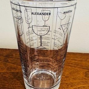 May include: Clear glass cocktail shaker with black printed cocktail recipes, including Margarita, Alexander, and Martini. The shaker has measurement markings in milliliters and ounces, and instructions for shaking with ice.