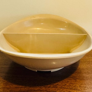 May include: A two-toned, oval-shaped serving bowl with a built-in divider. The bowl features a light tan interior and a white rim and base. The design is mid-century modern, suitable for serving snacks or small dishes. The bowl is sitting on a wooden surface.
