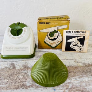 Può includere: Un set di affilacoltelli Dial-X vintage, con un affilacoltelli in plastica bianca e verde, un cono verde e una scatola con un'illustrazione di coltello. La scatola recita "CAP SNAFFLER SHARPENS KNIVES". Una piccola carta mostra l'affilacoltelli.