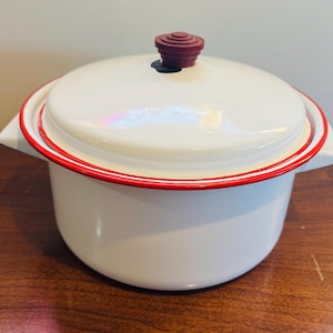 Könnte beinhalten: Weißer Emaille-Topf mit Deckel. Der Topf hat einen roten Rand um den Rand und einen dunkelroten Knopf auf dem Deckel. Der Topf hat zwei seitliche Griffe. Der Topf steht auf einer Holzoberfläche.