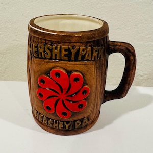 May include: A brown ceramic mug with the text "HERSHEYPARK" and a red flower design. The mug has a handle and a white interior. The mug is a souvenir from Hershey Park.
