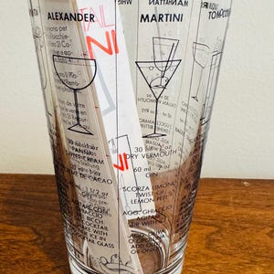 May include: A clear cocktail mixing glass with black printed cocktail recipes, including the Alexander and Martini. The glass has a flared shape and is filled with a white and red recipe card and a stirring rod.