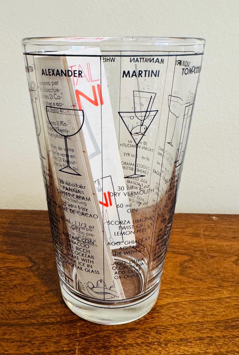 May include: A clear cocktail mixing glass with black printed cocktail recipes, including the Alexander and Martini. The glass has a flared shape and is filled with a white and red recipe card and a stirring rod.