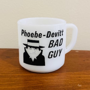 Può includere: Tazza in ceramica bianca con grafica nera di una persona con cappello e la scritta "Phoebe-Devitt BAD GUY". La tazza ha un manico curvo ed è appoggiata su una superficie di legno.