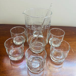 Puede incluir: Un juego de cóctel vintage de vidrio transparente, que incluye una jarra con un diseño punteado, un agitador y seis vasos bajos a juego. El juego está dispuesto sobre una superficie de madera.