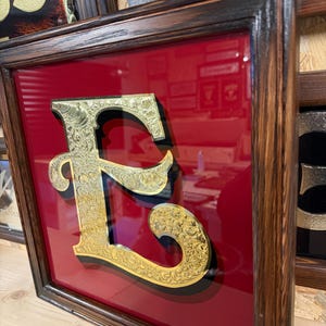 May include: Framed gold-colored letter "E" with ornate floral detailing, set against a red background. The letter is raised, creating a 3D effect. The frame is dark brown with a wood grain finish. Other framed letters are visible.