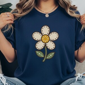 Puede incluir: Una camiseta de manga corta azul marino con un gráfico central de una flor color crema de cinco pétalos, un centro amarillo y un tallo verde con dos hojas. El diseño de la flor incluye contornos marrón oscuro y detalles de costura.