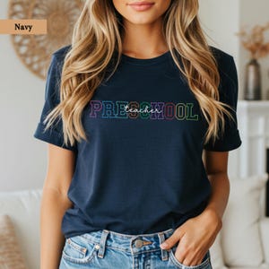 Puede incluir: Camiseta azul marino con la frase "Preschool teacher" en letras coloridas y delineadas. La camiseta es de cuello redondo y está hecha de un material suave. El texto es multicolor.