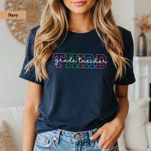 Puede incluir: Camiseta azul marino con la frase "THIRD grade teacher" en letras coloridas. La palabra "THIRD" está en rosa, verde, naranja y azul. El texto "grade teacher" está en un estilo informal y manuscrito.