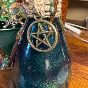 Peut inclure: Une bouteille en verre bleu-vert foncé avec un pendentif pentagramme doré. La bouteille a une finition scintillante et est ornée de corde et d'éléments décoratifs. Le design suggère un thème mystique ou fantastique.