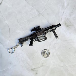 May include: A miniature black and silver gun keychain with a scope and a silver key ring. A quarter is next to the keychain for size comparison. The gun has a cylindrical magazine and a handle.