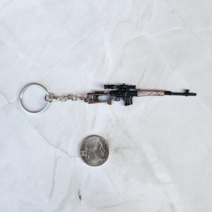 May include: Miniature black and brown rifle keychain with a scope, attached to a silver keyring. A quarter is next to the keychain for size comparison. The rifle has a detailed design.