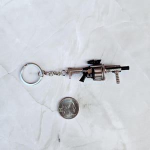 May include: Miniature silver-colored gun keychain with a black handle and scope. The keychain is attached to a silver chain and ring. A quarter is next to the keychain for size comparison.