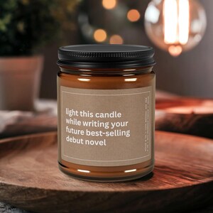 May include: A brown candle in a glass jar with a black lid. The label reads "light this candle while writing your future best-selling debut novel." The candle sits on a wooden tray.