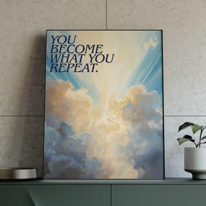 May include: A dark green modern sideboard with gold legs, holding a framed art print with the text "YOU BECOME WHAT YOU REPEAT." The print features a sky with clouds. A small potted plant and a lamp are also on the sideboard.