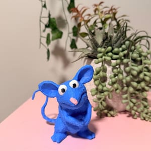 May include: A blue mouse figurine with large ears, white eyes, and a pink nose. The mouse is positioned on a pink surface, with a potted plant in the background. The figurine has a whimsical, cartoonish design.