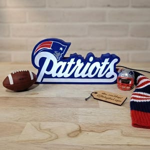 May include: A blue and white Patriots logo with a football, miniature helmet, and a red, white, and blue striped beanie. A wooden tag with text is also present. The items are arranged on a wooden surface.