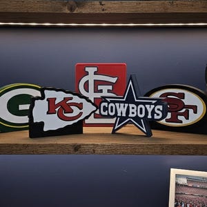 May include: A wooden shelf displays sports team logos and a pair of black headphones. The logos include the Green Bay Packers, Kansas City Chiefs, St. Louis Cardinals, Dallas Cowboys, and San Francisco 49ers. The shelf is made of wood and has a dark blue background.