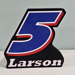 May include: A black, white, red, and blue number 5 sign with the name "Larson" in white text. The number 5 is blue with a white border and red outline. The sign is a decorative item.